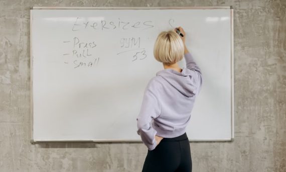 Female writing workout schedule on whiteboard in gym setting.